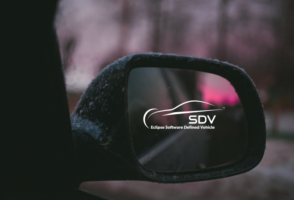 Close-up of a car’s side mirror on a cold day, with frost on the mirror housing. Reflected in the mirror is the ‘SDV – Eclipse Software Defined Vehicle’ logo, with blurred pink and white lights and trees in the background
