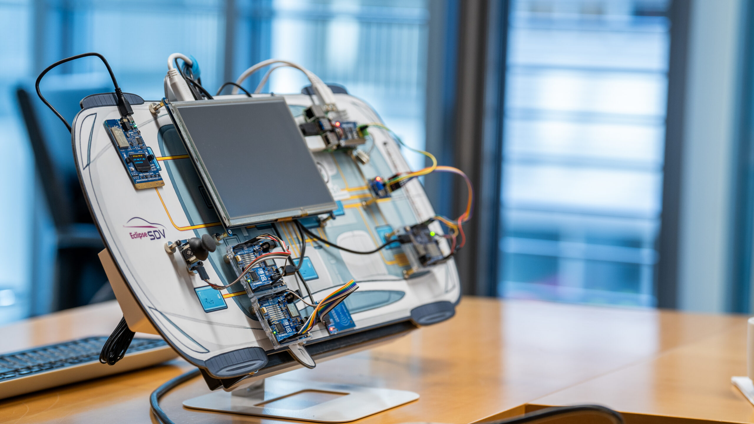 Prototype SDV development board mounted on a stand, featuring multiple connected microcontroller modules, wiring, and a central display, with an Eclipse SDV logo visible on the panel in an office setting.