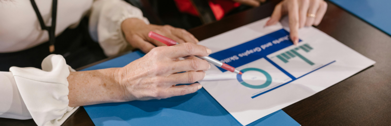 Close-up of two people reviewing a printed report with charts and graphs, one pointing at a bar chart while the other holds a pen, seated at a desk with folders.