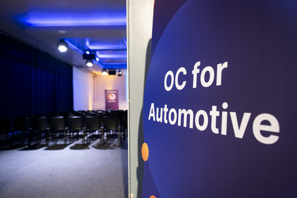 A blue sign with white letters saying "OC for Automotive", with an auditorium in the background.