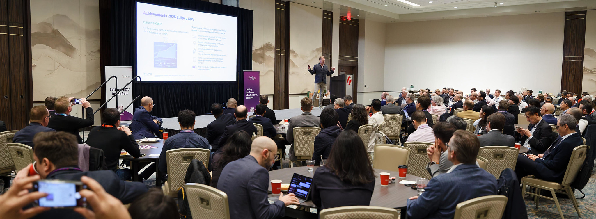 A large group of people sitting at tables in a big hotel conference room. In the back, a man standing on a stage, presenting a talk