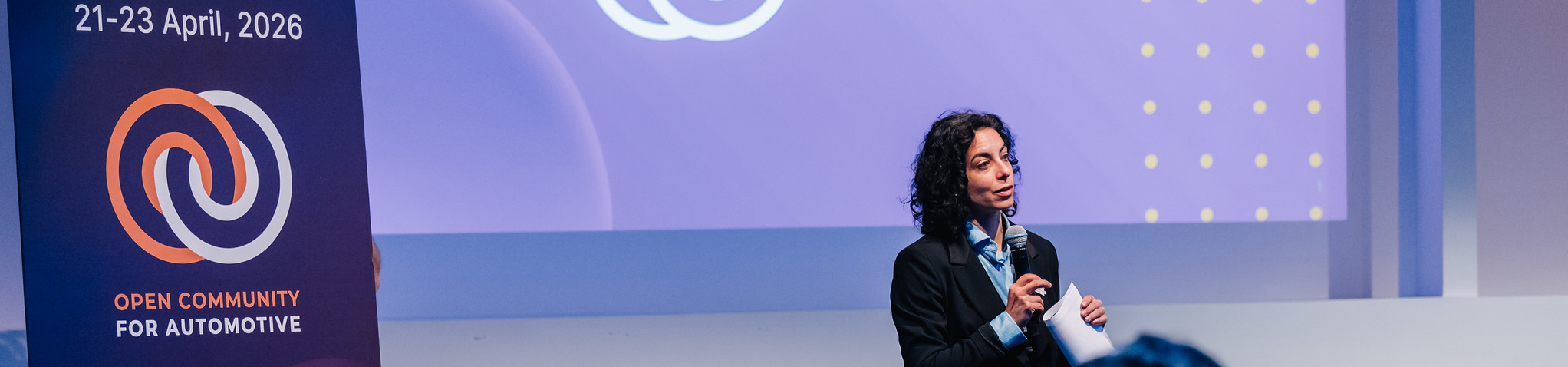 Woman with microphone on stage, standing next to a roll-up banner saying "Open Community for Automotive"